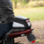 A motorcycle tail bag with a black, white, and gray color scheme and red accents, shown mounted on the back of a red motorcycle with a rider in a black leather jacket, set against a blurred outdoor background.