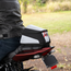 A motorcycle tail bag with a black, white, and gray color scheme and red accents, shown mounted on the back of a red motorcycle with a rider in a black leather jacket, set against a blurred outdoor background.