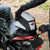 A black and white motorcycle tail bag with red zipper pulls is being mounted on a motorcycle tail light by a rider wearing black gloves. The helmet features a white logo on the side.