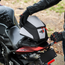 A black and white motorcycle tail bag with red zipper pulls is being mounted on a motorcycle tail light by a rider wearing black gloves. The helmet features a white logo on the side.