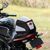 A motorcycle tail bag in black, white, and gray with red accents, shown mounted on the rear of a black motorcycle in an outdoor grassy setting.