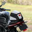 A motorcycle tail bag in black, white, and gray with red accents, shown mounted on the rear of a black motorcycle in an outdoor grassy setting.